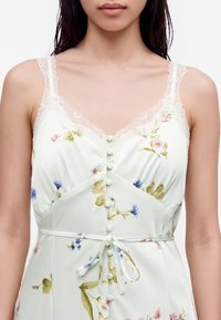 Γυναίκα που φοράει λευκό φόρεμα με floral σχέδιο, δαντελένιους ιμάντες και κουμπιά μπροστά, δεμένο με λεπτή ζώνη στη μέση.