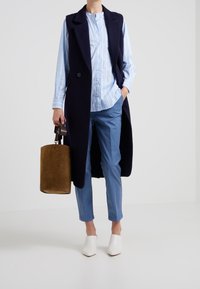 Personne portant un manteau sans manches bleu marine par-dessus une chemise à rayures bleu clair et un pantalon bleu, tenant un sac à main cylindrique marron et des chaussures blanches pointues.