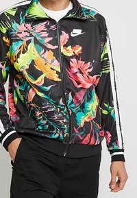 Svart jacka med livfulla blommönster i rosa, gult och grönt. Har dragkedjestängning, vita detaljer och en Nike-logotyp på bröstet.