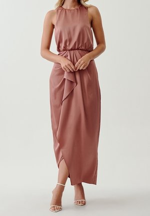 Femme portant une robe longue sans manches couleur rose poussiéreux avec une taille froncée et une jupe drapée asymétrique, associée à des talons blancs à brides.