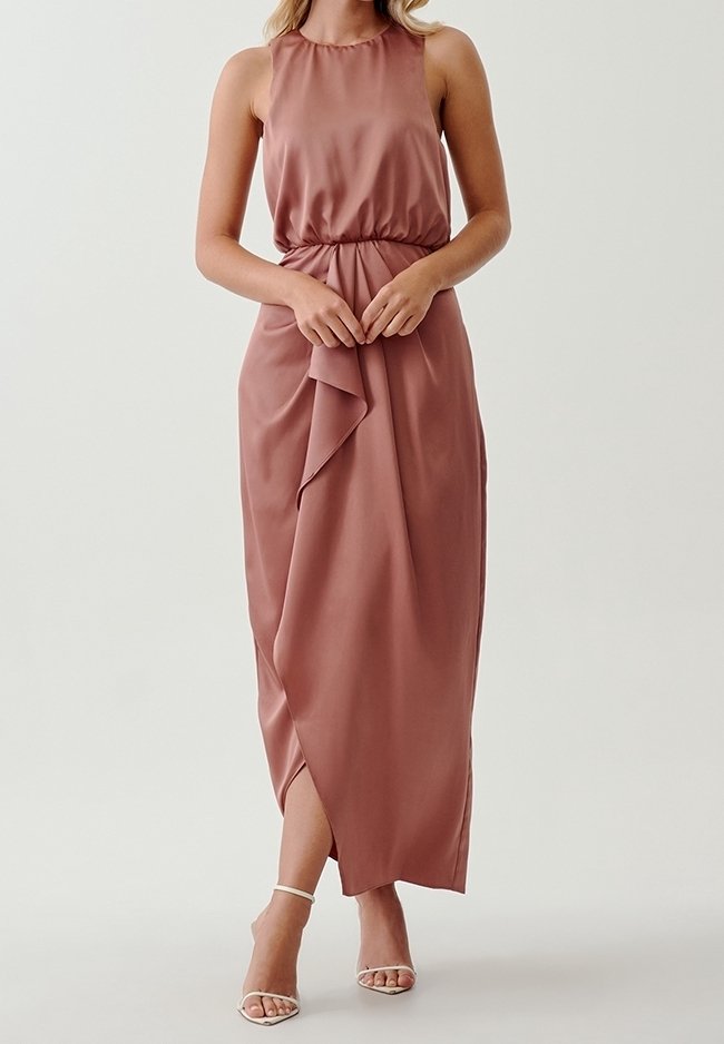 Femme portant une robe longue sans manches couleur rose poussiéreux avec une taille froncée et une jupe drapée asymétrique, associée à des talons blancs à brides.