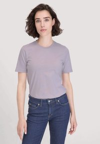 Jonge vrouw met kort krullend haar, gekleed in een lichtpaars T-shirt en donkerblauwe jeans, staande tegen een effen lichte achtergrond.