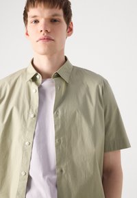 Camisa de manga corta, de color verde claro, con cuello puntiagudo, cierre frontal de botones y textura suave, superpuesta sobre una camisa blanca.