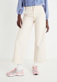 Pantalones de pierna ancha beige, de tela de algodón, de talle alto, con cierre de botón; combinados con zapatillas deportivas rosas y grises y calcetines blancos.