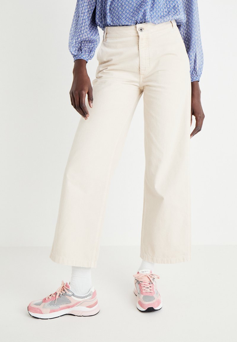 Pantalones de pierna ancha beige, de tela de algodón, de talle alto, con cierre de botón; combinados con zapatillas deportivas rosas y grises y calcetines blancos.