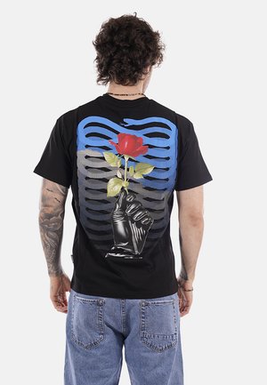 Uomo con capelli ricci che indossa una maglietta nera con una grafica di una mano guantata di nero che tiene una rosa rossa su sfondo a strisce blu e nere.