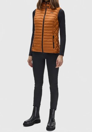 Personne portant un gilet matelassé sans manches orange par-dessus un haut noir à manches longues, un pantalon noir et des bottines noires, debout sur un fond uni.
