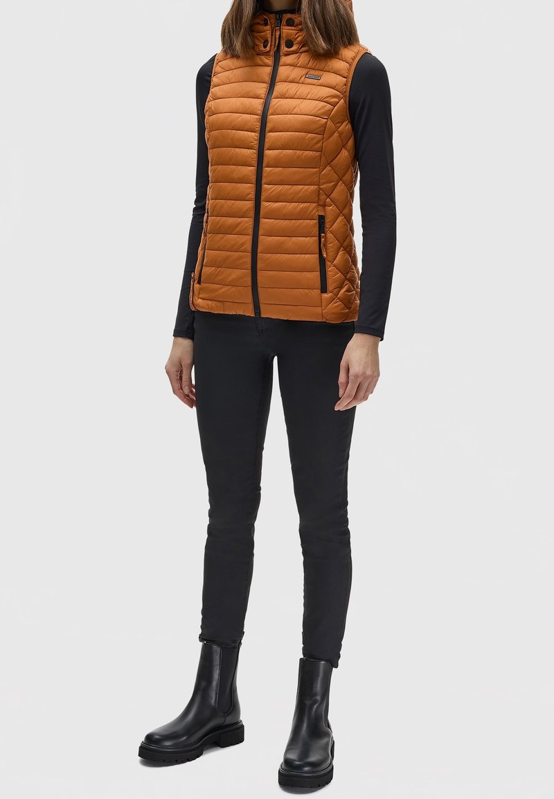 Personne portant un gilet matelassé sans manches orange par-dessus un haut noir à manches longues, un pantalon noir et des bottines noires, debout sur un fond uni.