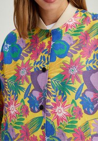 Kobieta nosząca żółtą kurtkę z jasnoniebieskimi, różowymi, fioletowymi i zielonymi tropikalnymi wzorami kwiatowymi oraz czarnymi zatrzaskami.