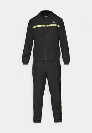 Hanorac negru cu fermoar complet și set de pantaloni de trening negri asortați, cu dungi orizontale galbene și logo Lacoste pe ambele piese.