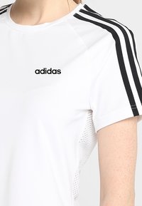 Vit atletisk t-shirt med svart Adidas-logotyp och tre svarta ränder på axlarna. Tillverkad med ventilerande mesh-paneler för luftgenomströmning.