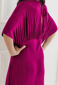 Robe fuchsia avec un détail noué dans le dos, manches courtes larges, taille froncée et finition lisse texturée.
