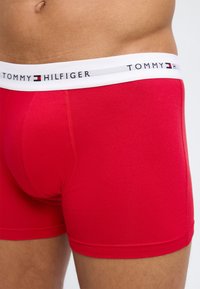 Männlicher Oberkörper trägt rote Tommy Hilfiger Boxershorts mit weißem Bund und Logo vor schlichtem Hintergrund.