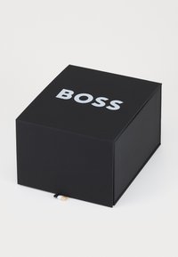 Boîte rectangulaire noire avec le mot "BOSS" en grandes lettres blanches sur le dessus. Présente une petite accent noir, blanc et or sur le côté.