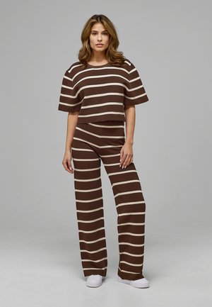 Femme debout portant un top à manches courtes rayé horizontalement marron et beige et un pantalon large assorti avec des baskets blanches sur fond gris.