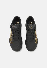 Sneaker nera con superficie texturizzata e design ricamato oro. Presenta lacci piatti, colletto imbottito e dettagli perforati nell'area delle punte.