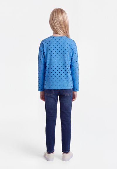 Barn med langt blondt hår iført blå langermet skjorte med polka prikker, mørkeblå jeans og hvite joggesko, sett bakfra.