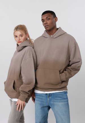 Δύο ενήλικες που φοράνε μπεζ φούτερ με κουκούλα, στέκονται δίπλα-δίπλα μπροστά σε ένα απλό ανοιχτόχρωμο φόντο, με χαλαρή έκφραση.