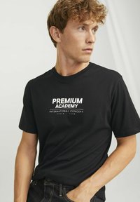 Črna bombažna majica s krožnim izrezom, z belim natisnjenim besedilom: "PREMIUM ACADEMY INTERNATIONAL CONCEPT SINCE 1990." Kratke rokave.