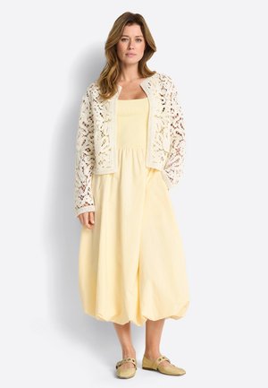 Woman wearing pale yellow midi dress, cream crochet lace open cardigan, and matching yellow flats with ankle straps, standing against white background.