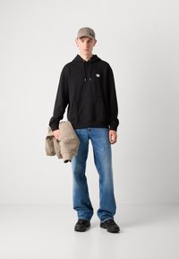 Zwarte hoodie met een voorzak, gecombineerd met lichtblauwe jeans. Model houdt een beige jack vast, draagt een beige pet en zwarte sneakers.