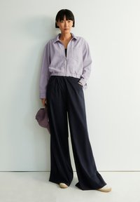 Chaqueta de cremallera lavanda con un bolsillo en la parte frontal combinada con pantalones anchos de color navy. El modelo sostiene una gorra de color púrpura claro, de pie sobre un suelo claro.