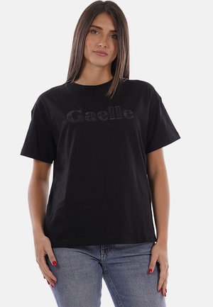 Donna con capelli castani lisci, indossa una t-shirt nera con il logo "Gaelle" e jeans blu, in piedi con le mani rilassate ai lati.