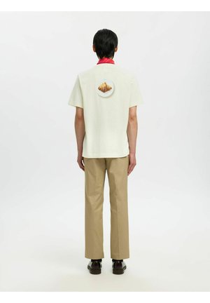 Osoba ubrana w kremową koszulkę z krótkim rękawem z nadrukiem gór na plecach, czerwoną ozdobą na kołnierzyku, beżowymi spodniami i czarnymi butami, stojąca tyłem.