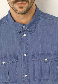 Camicia in denim blu con colletto a bottoni, due tasche frontali con pattina e bottoni a pressione metallici per la chiusura. Tessuto morbido e con texture.