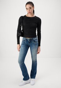 Black long-sleeve top with gathered front detail, paired with blue distressed bootcut jeans and white sneakers. Accessorized with a small black bag.