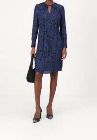 Långärmad klänning i marinblått med blå blomsterdesign, midjeband, V-ringning och en något utsvängd nederdel. I kombination med en svart vävd väska och klackar.