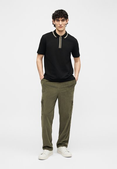 Νέος με σγουρά μαλλιά που φοράει μαύρο πόλο με κοντά μανίκια, χακί παντελόνι cargo, λευκά αθλητικά παπούτσια, στέκεται με τα χέρια στις τσέπες.