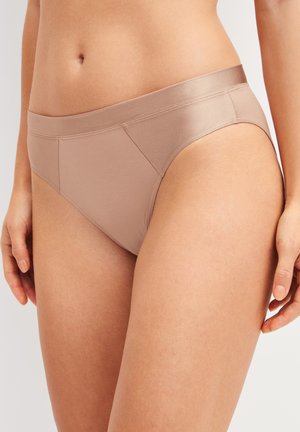 Femme portant des sous-vêtements beiges taille haute, montrée de la taille jusqu'au milieu de la cuisse, les mains détendues sur les côtés sur un fond neutre.