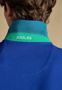 Gros plan sur le cou et le dos d'une personne montrant un col polo bleu et vert avec "JOULES" brodé sur la partie verte.