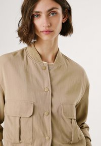 Beige bomber jacket made of smooth fabric, featuring a ribbed collar, two front pockets, and snap button closures.