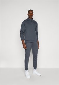 Grå hettegenser med høy krage og brodert logo, sammen med matchende bukser med lommer og ribbestrikkede mansjetter. Hvite sneakers.