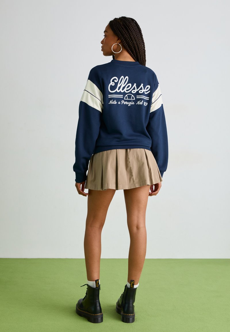 Sweatshirt bleu marine avec des lettres blanches et des rayures crème ; associé à une jupe beige plissée et des bottines noires à lacets. Fond de sol vert.