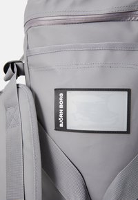 Sac à dos gris avec une surface texturée, des bretelles réglables, une poche zippée à l'avant et un porte-nom en plastique avec un cadre noir.