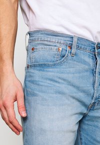 Los jeans de mezclilla azul claro presentan un diseño clásico y entallado con acentos de botones de cobre y un desvanecimiento distintivo. Incluyen bolsillos frontales y presillas para el cinturón.