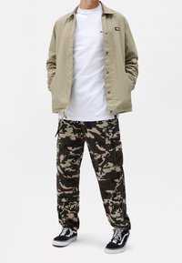 Ljusbeige knappjacka, vit t-shirt, kamouflage cargo-byxor med fickor och svarta sneakers med vita sulor. Slät tyg, avslappnad passform.