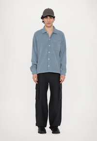 Light blue corduroy shirt with a chest pocket, paired with black cargo pants featuring side pockets, and black sneakers. Gray cap on head.