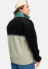 Corduroy and fleece jacket in black, green, and beige. Features a high collar and patterned trim. Relaxed fit with a textured finish.