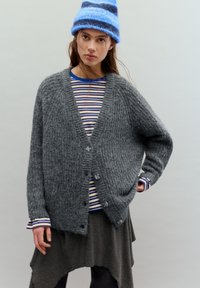 Cardigan gri tricotat cu material texturat, cu decolteu adânc în V și patru nasturi. Dedesubt, o cămașă în dungi cu tonuri albastre și albe.