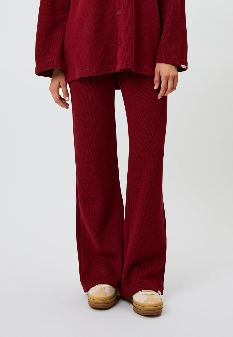 Bordeaux loungestel met een ribbelovershirt met knopen en bijpassende wijde broek. Zachte textuur, ontspannen pasvorm, gecombineerd met beige sneakers.