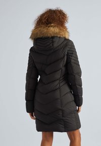 Schwarzer Steppmantel mit Chevron-Muster, ausgestattet mit einer abnehmbaren braunen Pelzkapuze und langen Ärmeln, reicht bis zur Mitte der Oberschenkel.