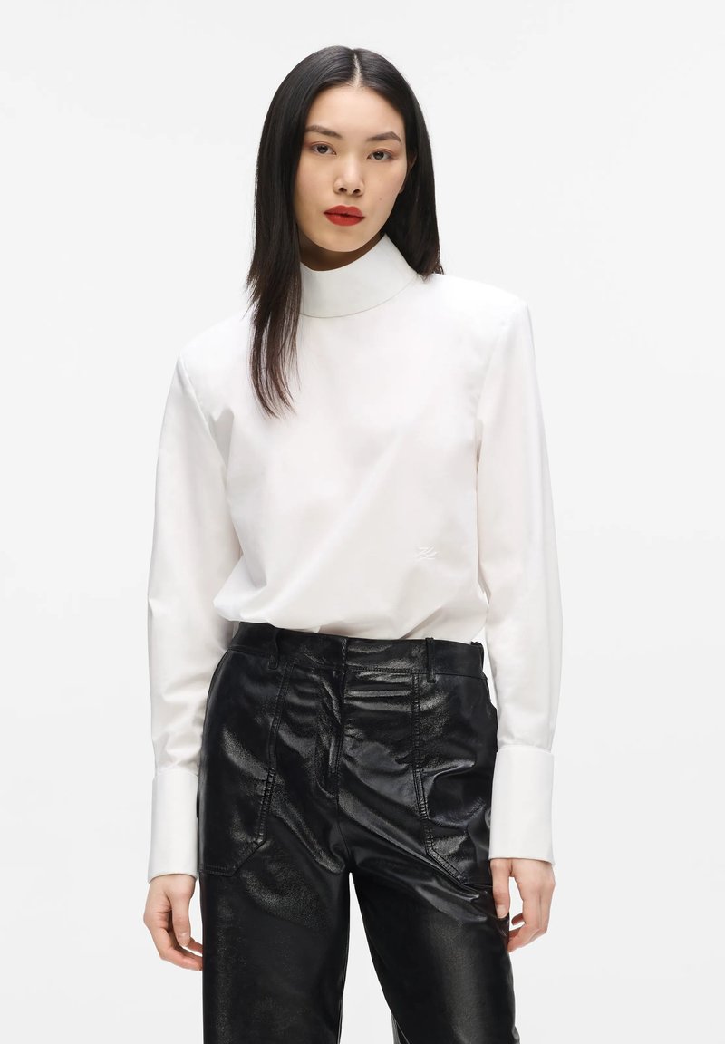 Femme aux cheveux noirs et raides porte un chemisier blanc à col montant et un pantalon en cuir noir brillant sur un fond blanc uni.