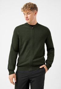 Młody mężczyzna z jasnymi kręconymi włosami, ubrany w ciemnozielony rozpinany sweter i czarne spodnie, stoi z jedną ręką w kieszeni.