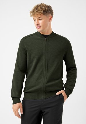 Junger Mann mit hellen, lockigen Haaren, der einen dunkelgrünen Reißverschlusspullover und schwarze Hosen trägt und mit einer Hand in der Tasche steht.