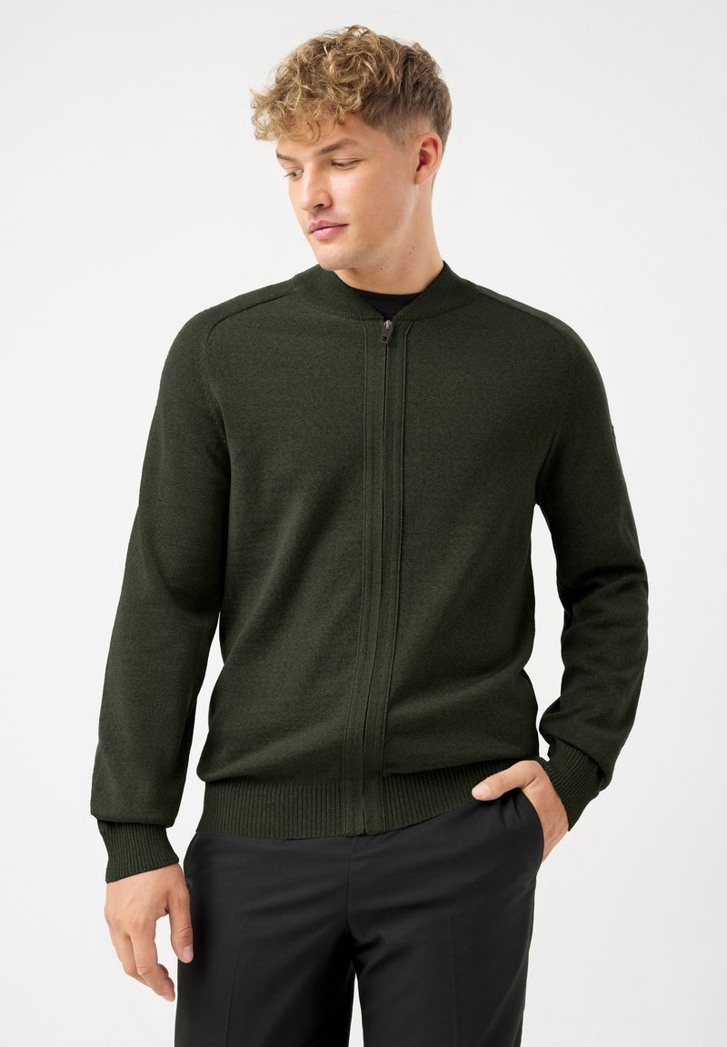 Młody mężczyzna z jasnymi kręconymi włosami, ubrany w ciemnozielony rozpinany sweter i czarne spodnie, stoi z jedną ręką w kieszeni.