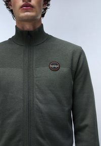 Kaki zip-up tröja med hög krage, texturerad överdel och en cirkulär logotypapplikation. Ribbad accent längs dragkedjan. Mjukt stickat material.